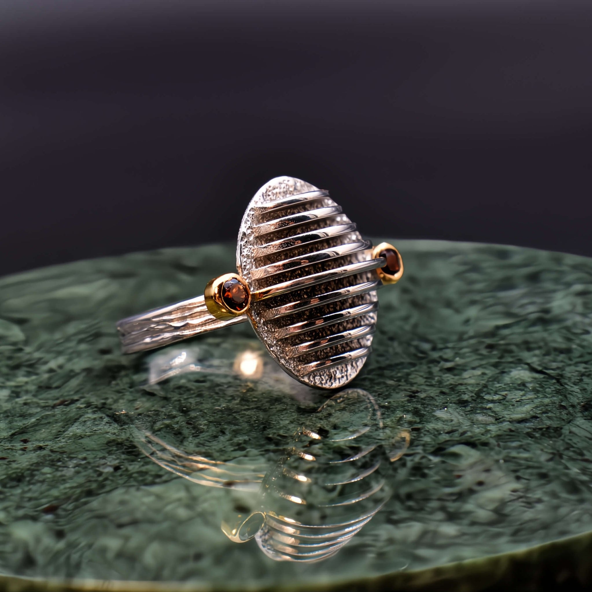 A sterling silver ring inspired by sand dunes, with a minimalistic, sculptural design. The ring features two gold plated bezels, each holding a small, deep red garnet stone on either side of the band. The silver surface has smooth, wave-like curves, capturing the essence of natural sand formations."

